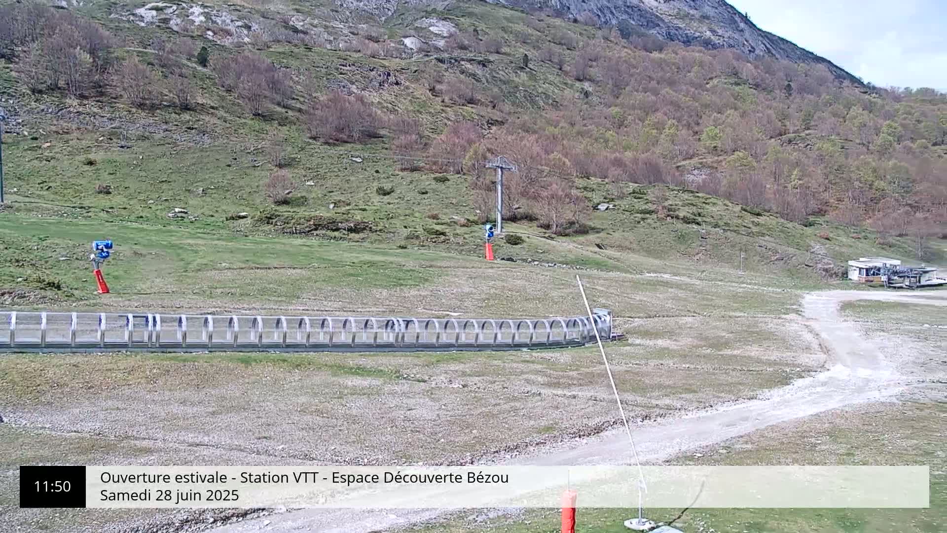 A mountainside view shows a grassy area with a curved track enclosed by a metal barrier, a chairlift, and a dirt road under a partly cloudy sky.