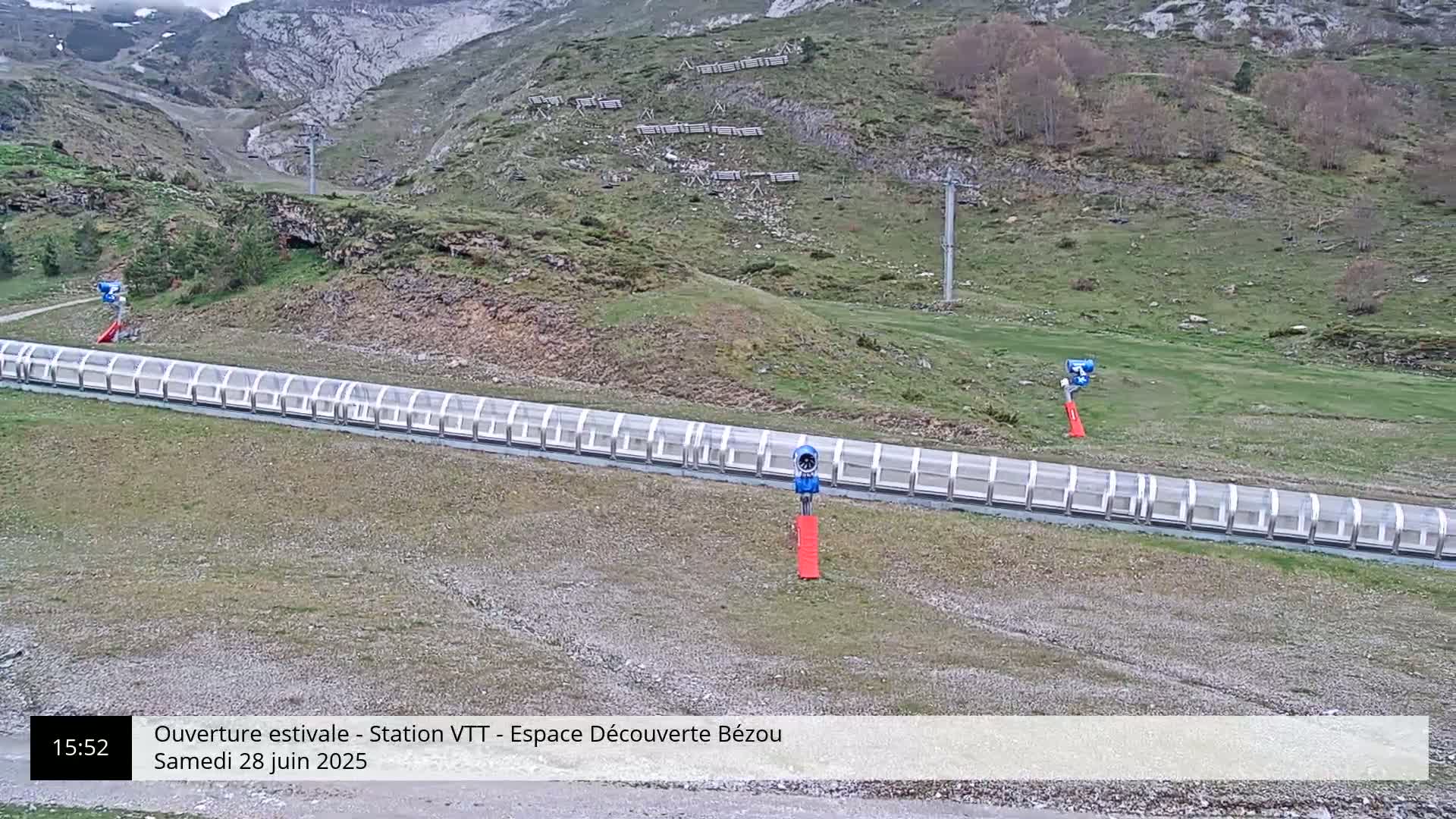 A long, covered conveyor belt is situated on a grassy hillside, with some sparse vegetation and a chairlift visible in the background under a partly cloudy sky.