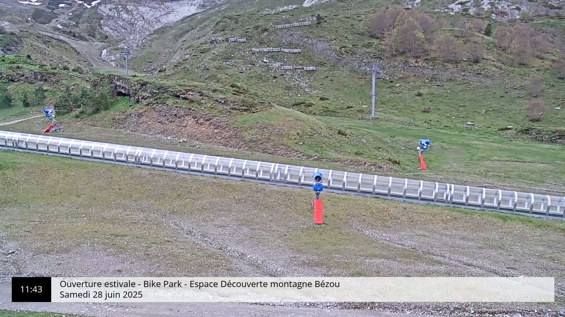 A long, covered conveyor belt sits in a grassy mountain area with a chairlift in the background on a partly cloudy day.