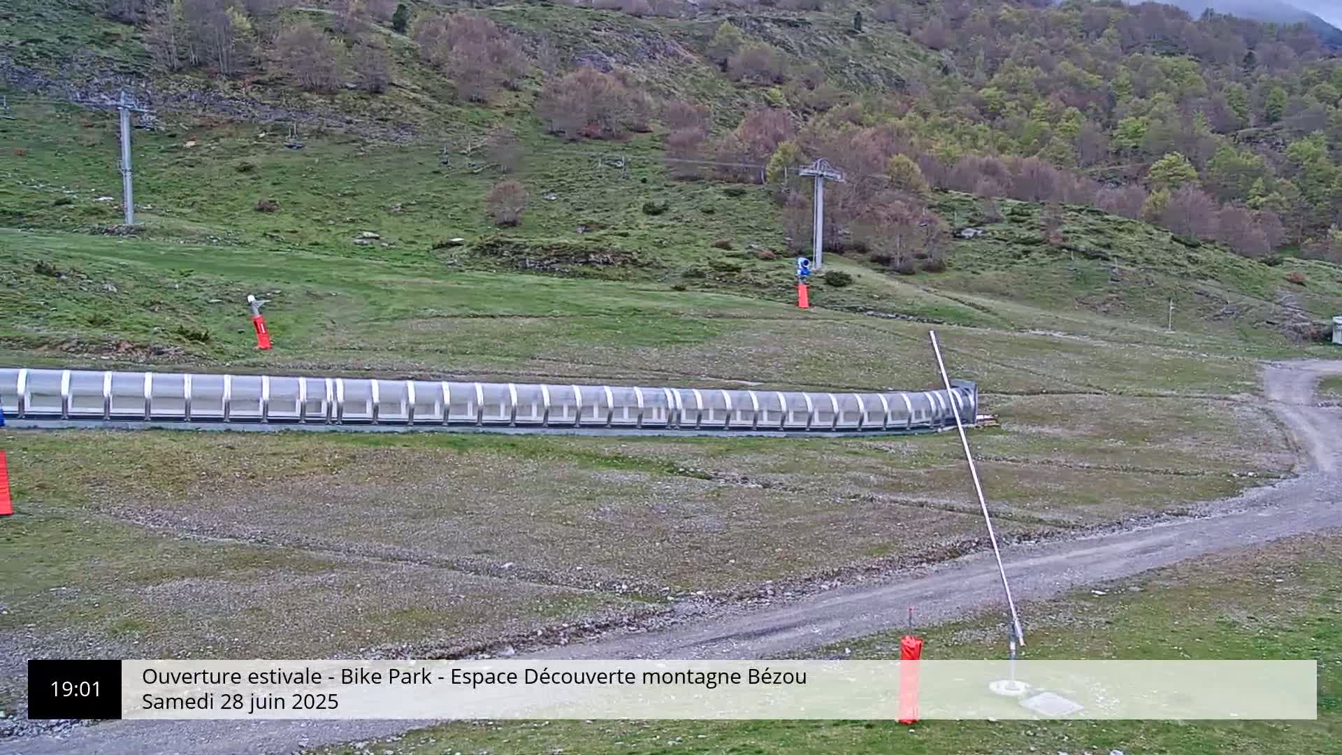 A long, covered track sits on a grassy hillside, with a chairlift visible in the background under a partly cloudy sky.