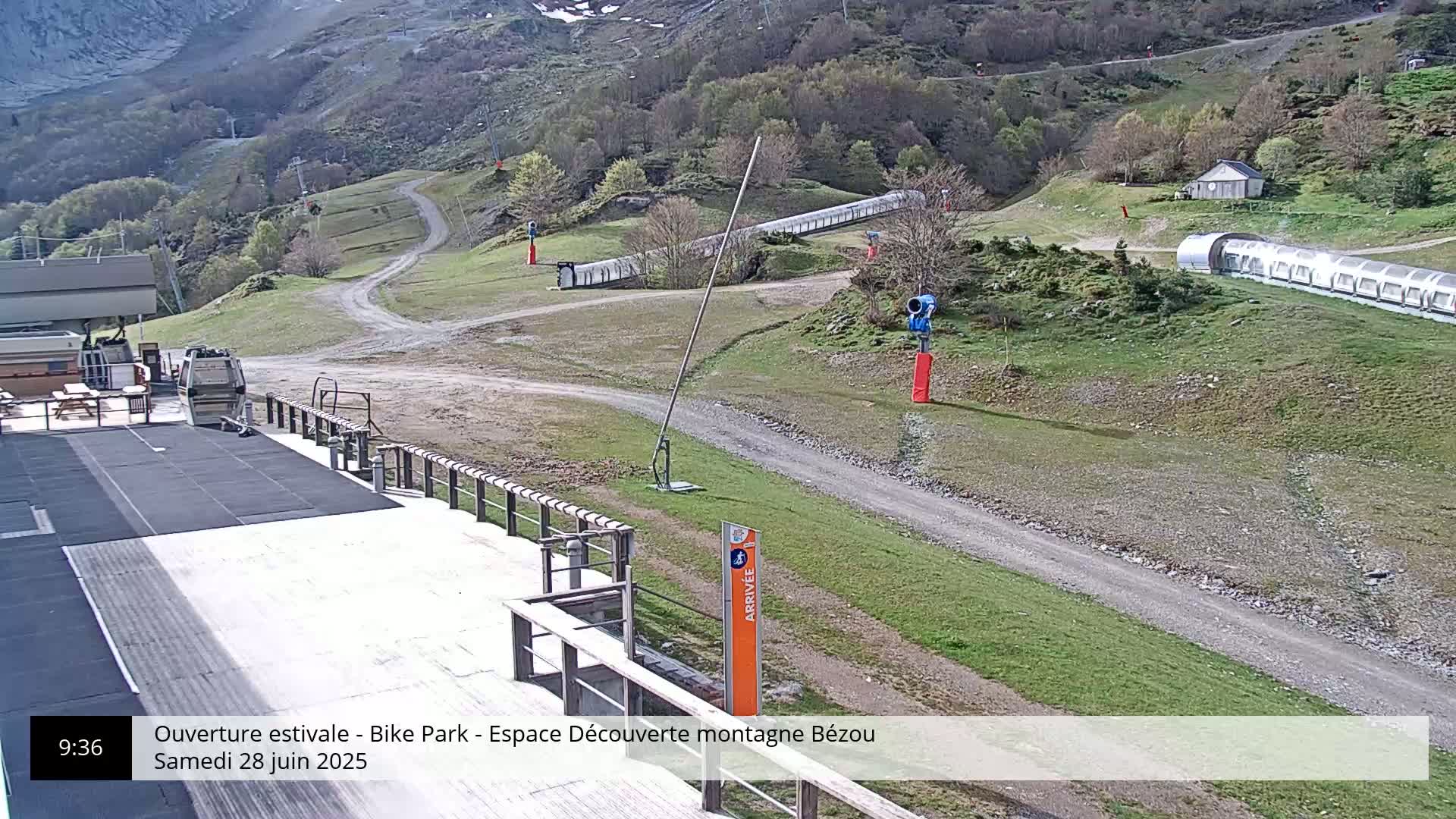 A mountain bike park with a dirt path, grassy areas, and snowmaking equipment, on a sunny day.