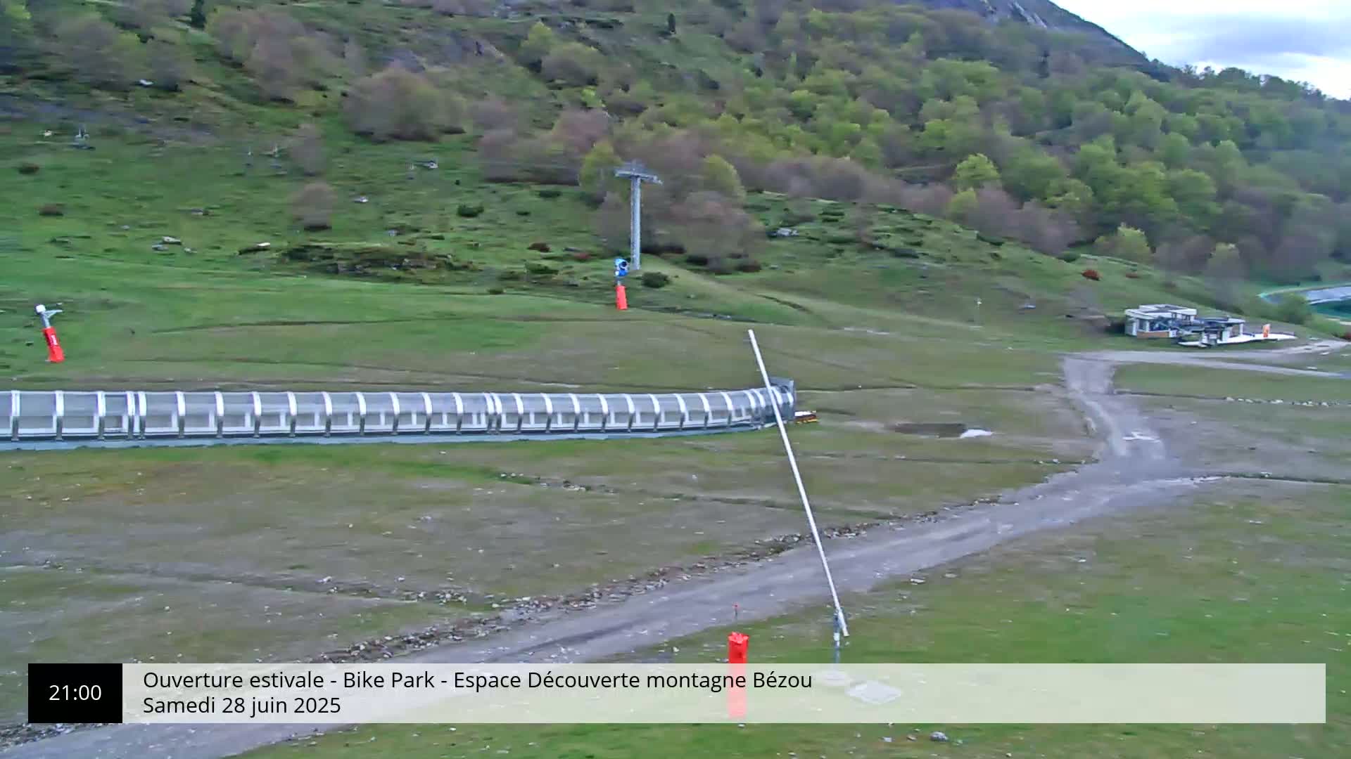 A grassy hillside with a ski lift, a covered track, and a small building is visible under an overcast sky.