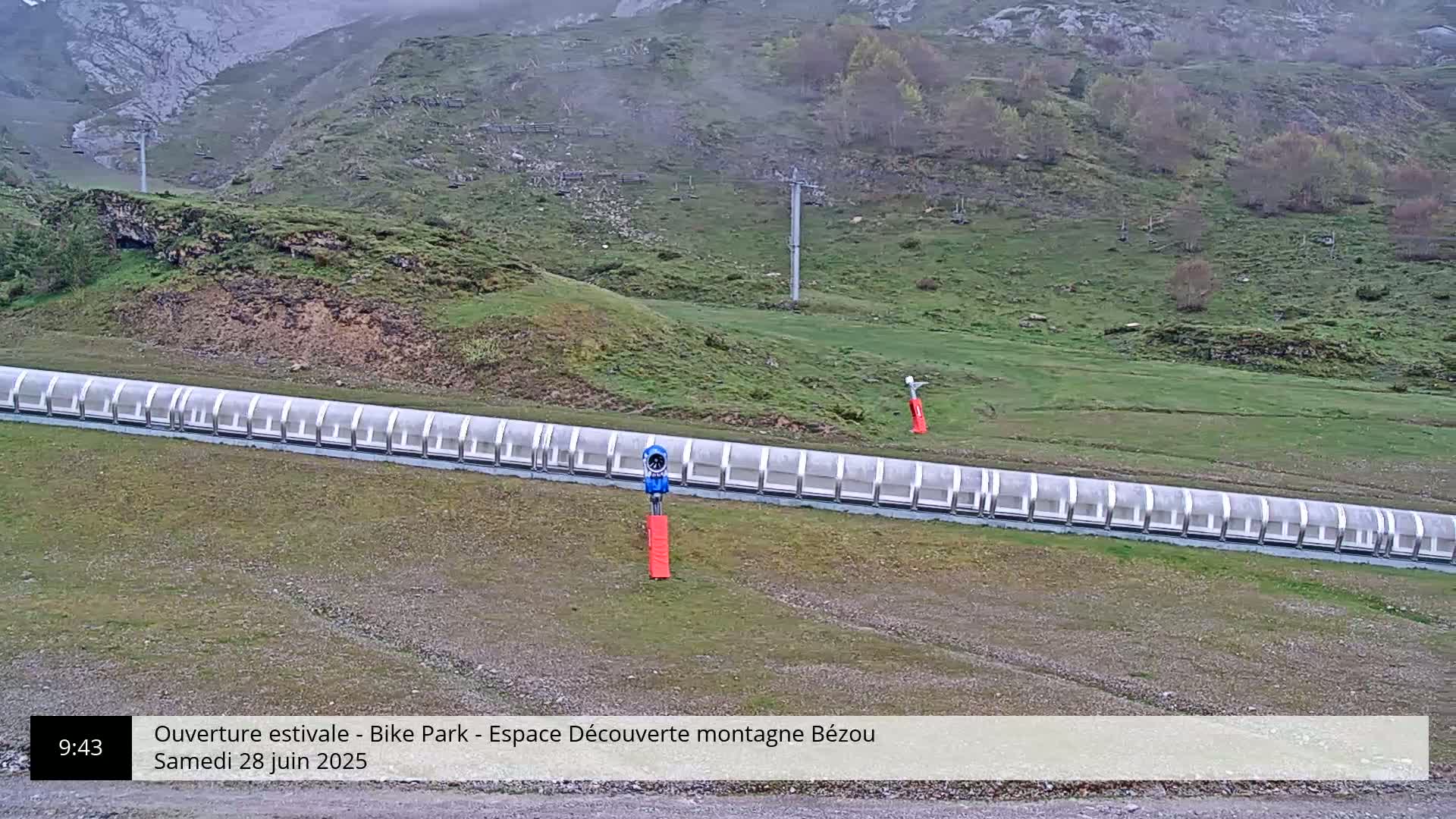 A long, covered conveyor belt sits on a grassy hillside against a mountainous backdrop under overcast skies.