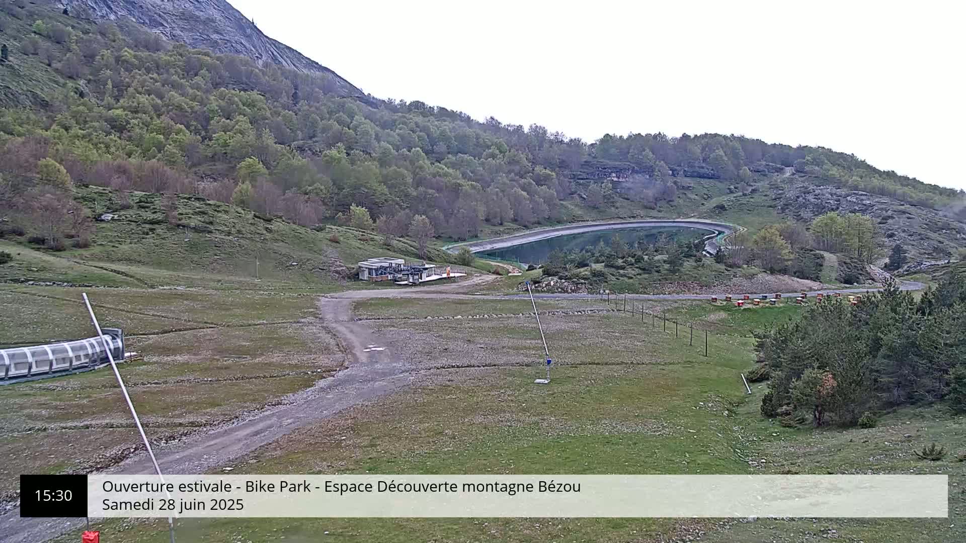 A mountainside view shows a dirt road leading to a small building near a calm body of water under an overcast sky.