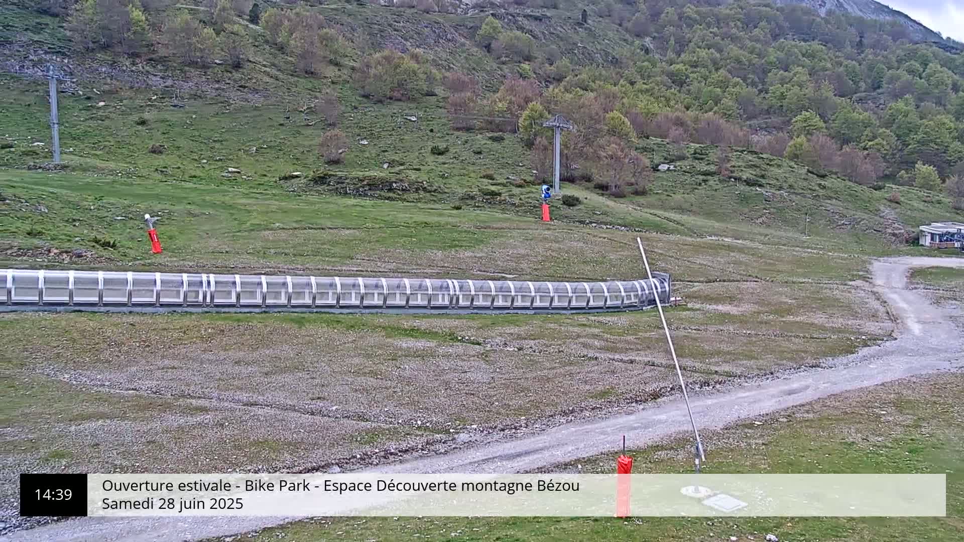 A long, covered track sits in a grassy area below a hillside with a chairlift and sparse trees, under sunny skies.