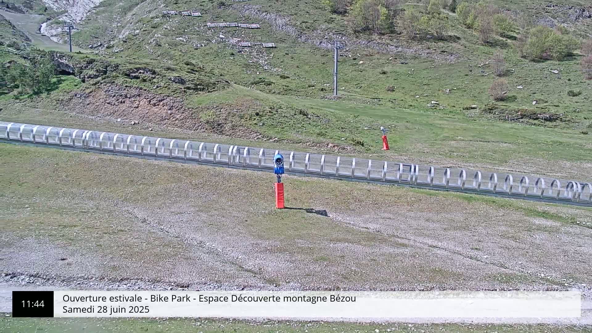A long, covered track stretches across a grassy hillside, with a blue snowmaking machine positioned near a red marker.