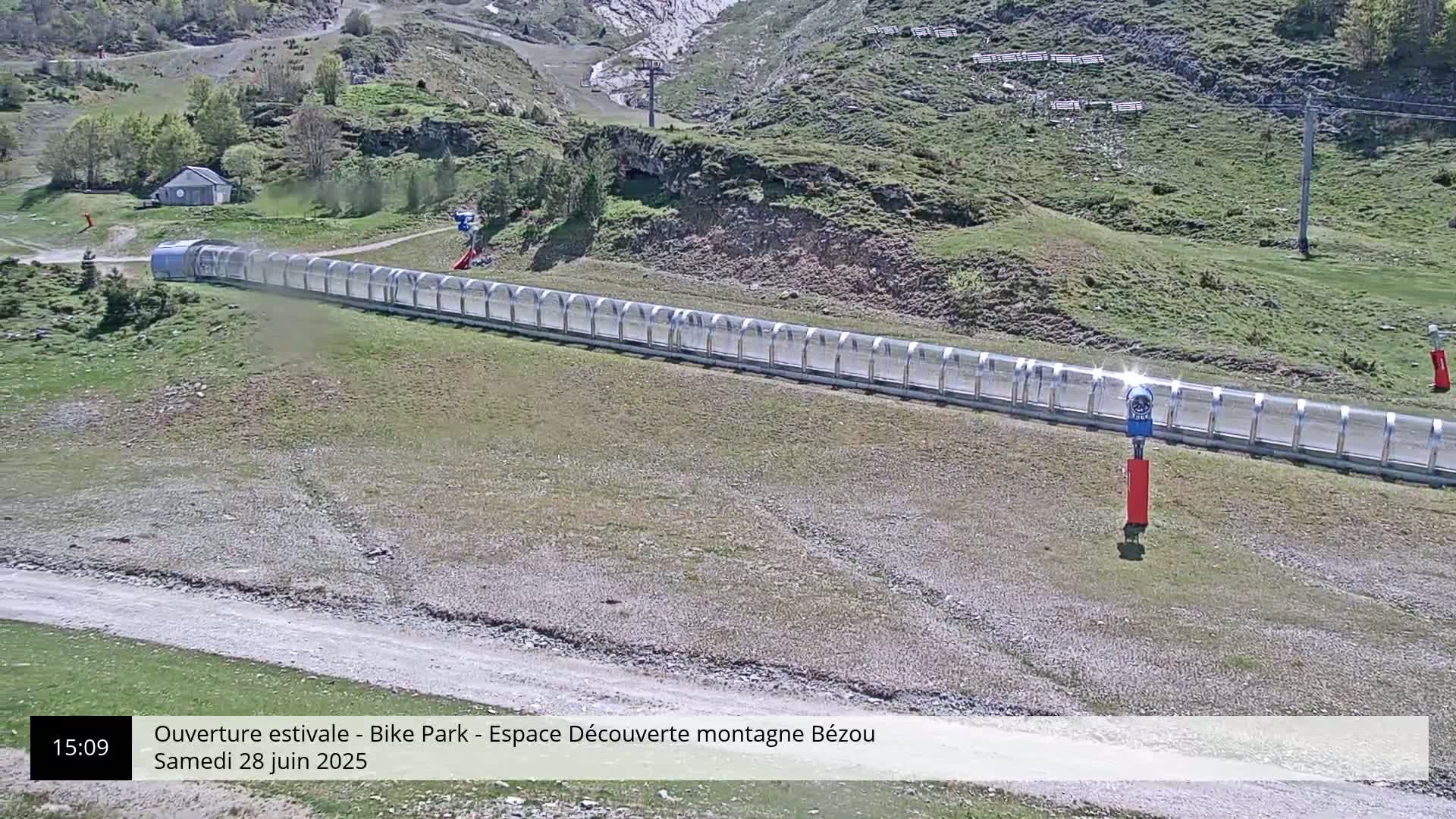 A long, covered track extends across a grassy hillside on a sunny day in a mountainous area.