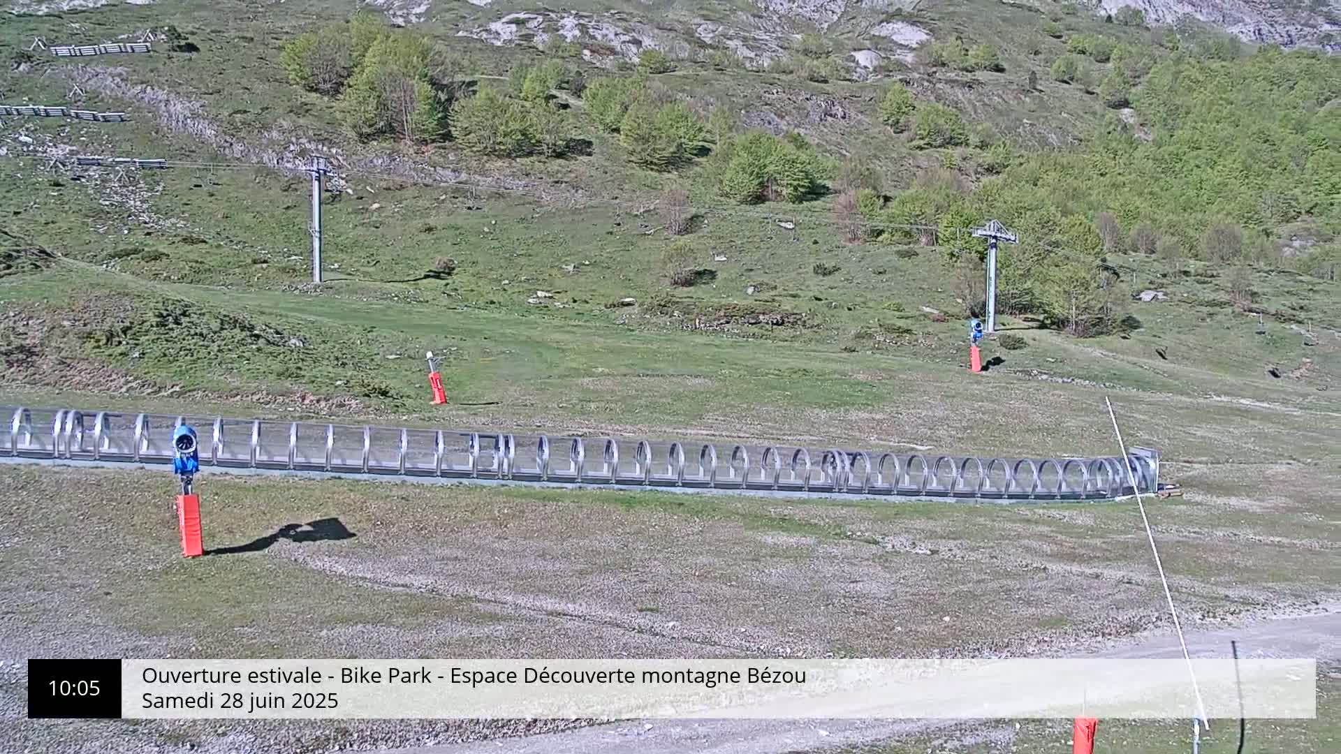 A long, covered structure sits on a grassy hillside below a ski lift, on a sunny day.