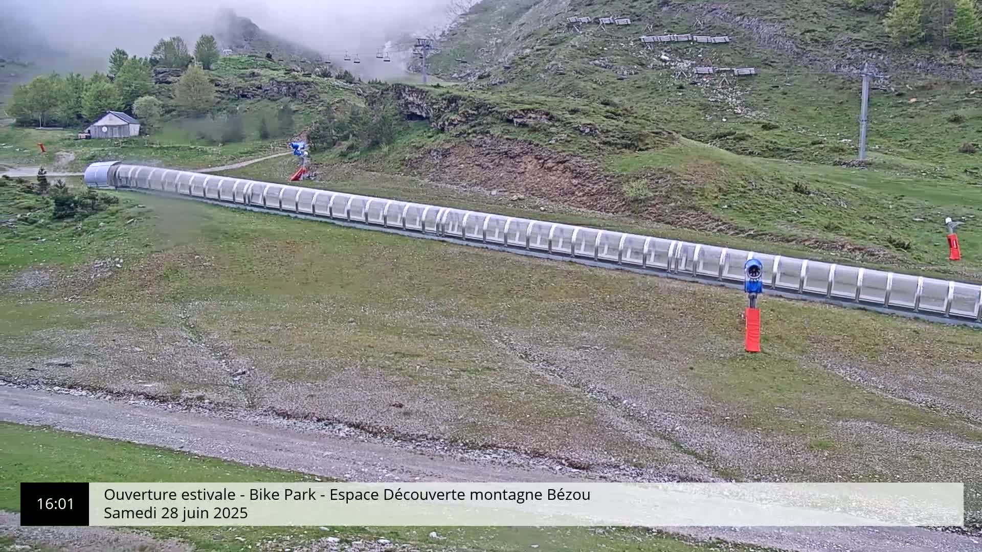 A long, covered track winds down a grassy hillside on a foggy day, with a chairlift visible in the background.