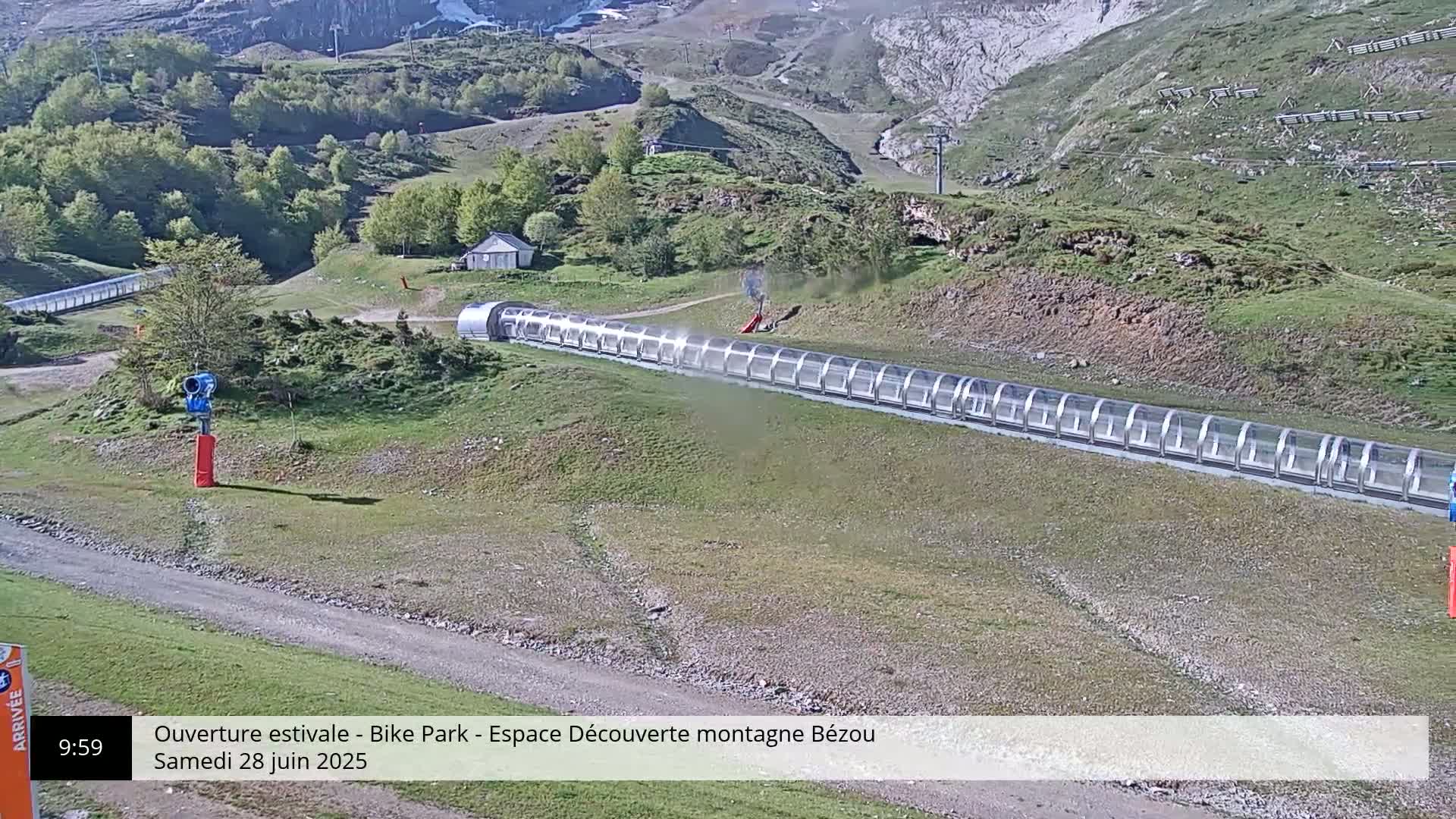 A long, covered conveyor belt sits on a grassy hillside under a sunny sky, with a snow-capped mountain in the background.