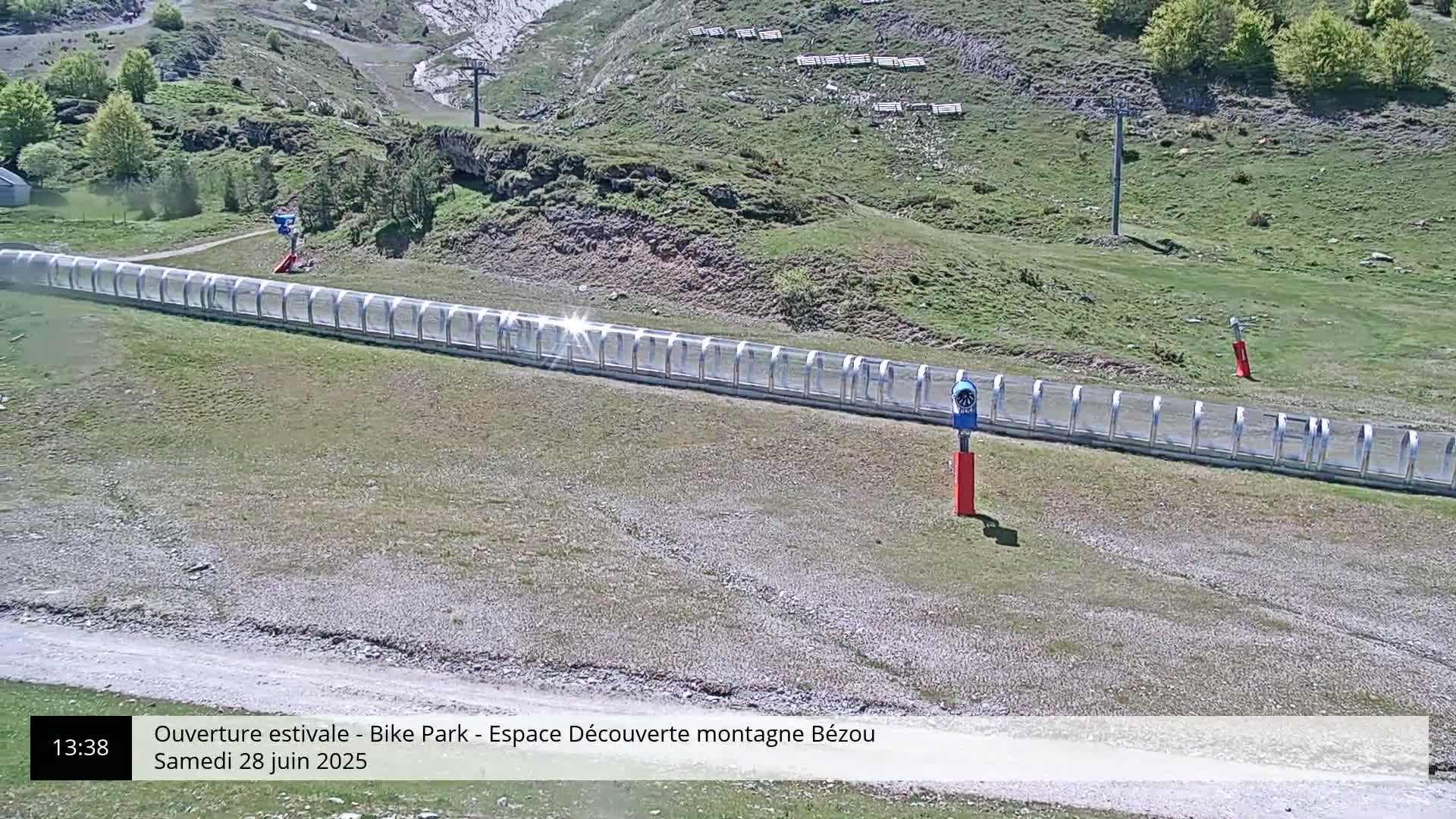 A long, transparent enclosure sits on a grassy hillside under a sunny sky, near a ski lift.