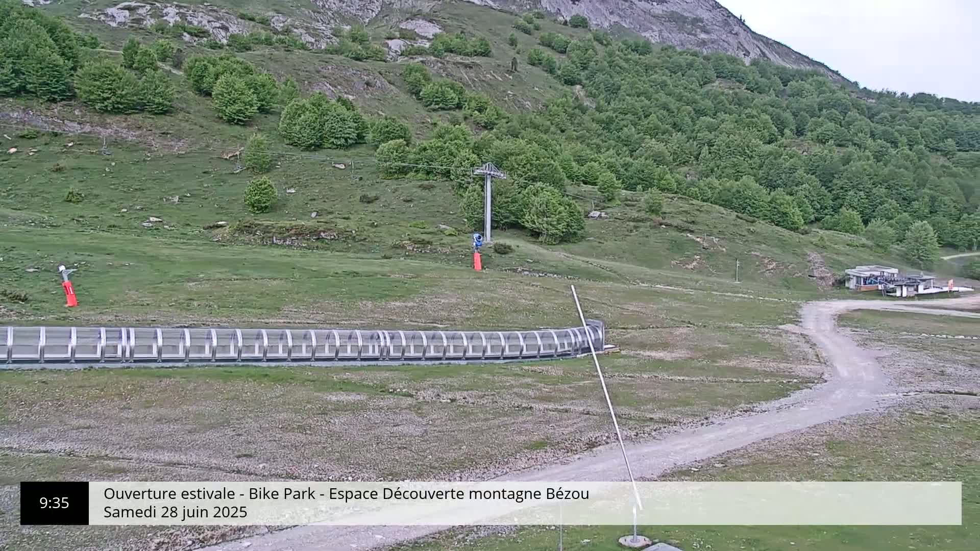A mountainside bike park with a covered track, a chairlift, and a small building is visible under an overcast sky.
