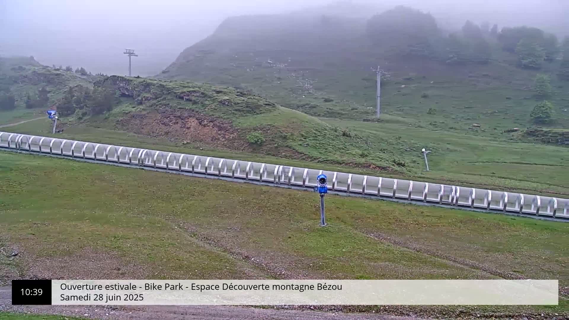 A long, covered track sits on a grassy hillside in foggy conditions, with a ski lift visible in the background.