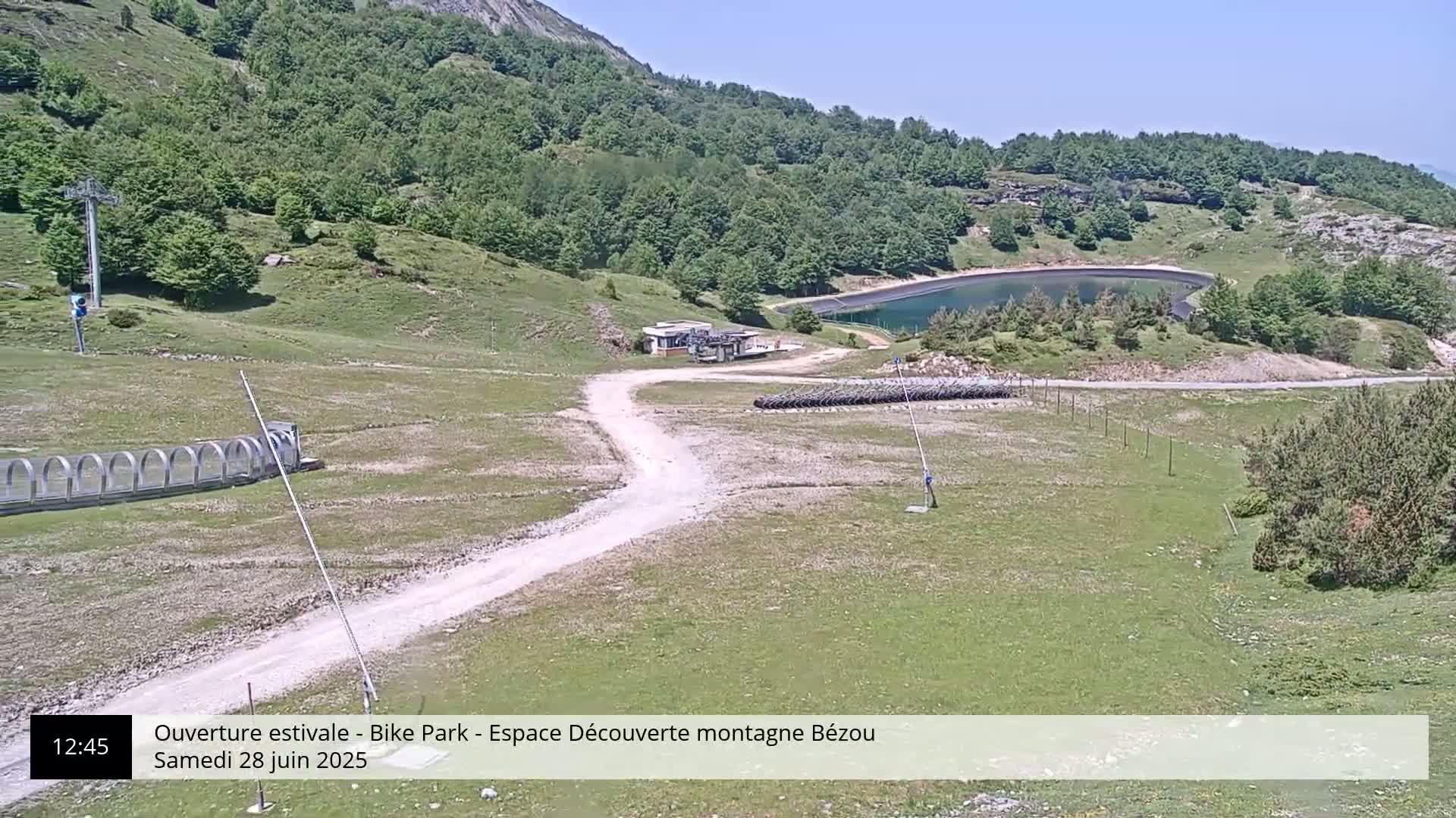 A mountaintop view on a sunny day shows a dirt road leading to a small building near a reservoir, with a grassy hillside and trees in the background.