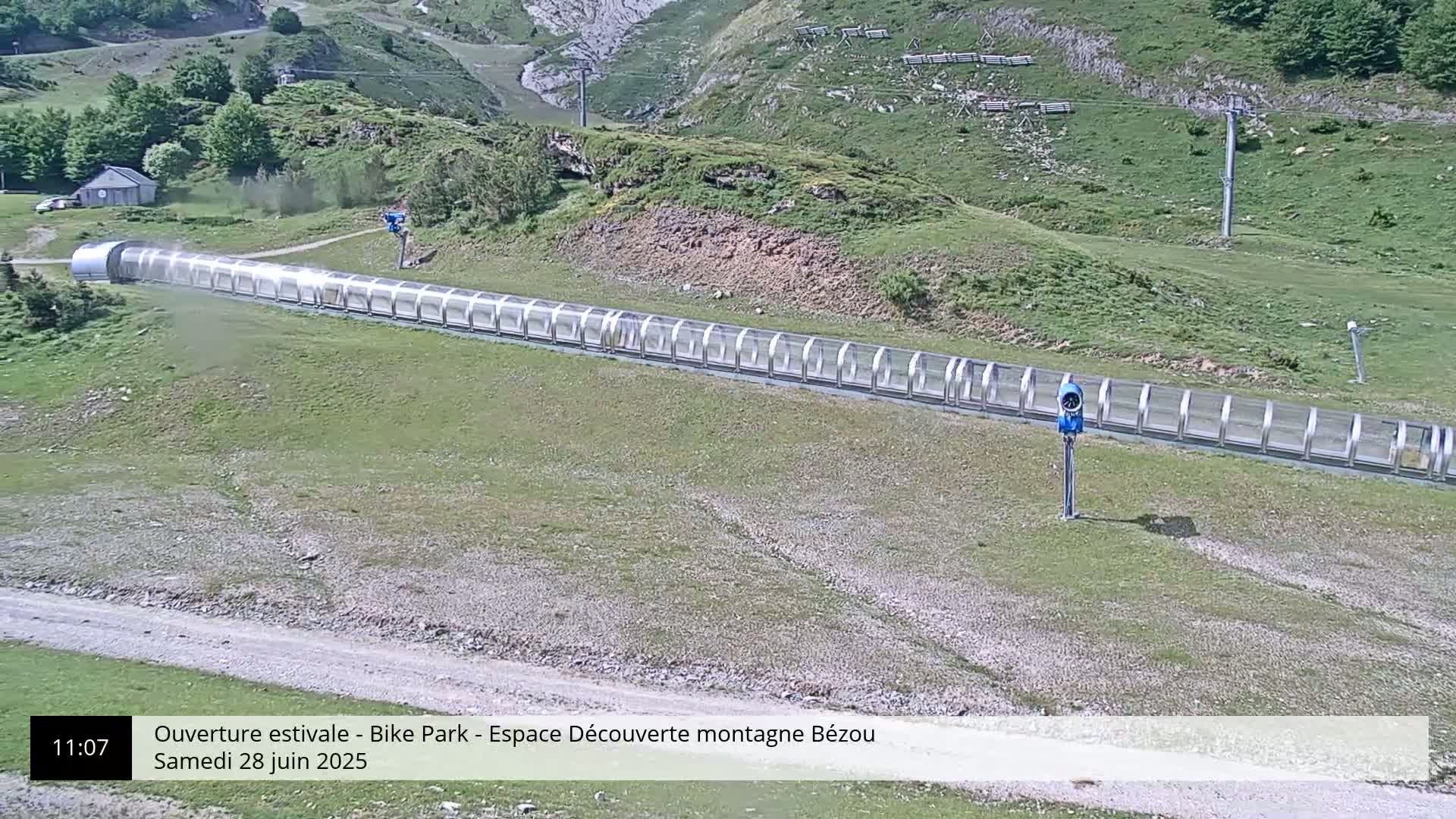A long, covered conveyor belt-like structure extends across a grassy hillside under a clear sky.