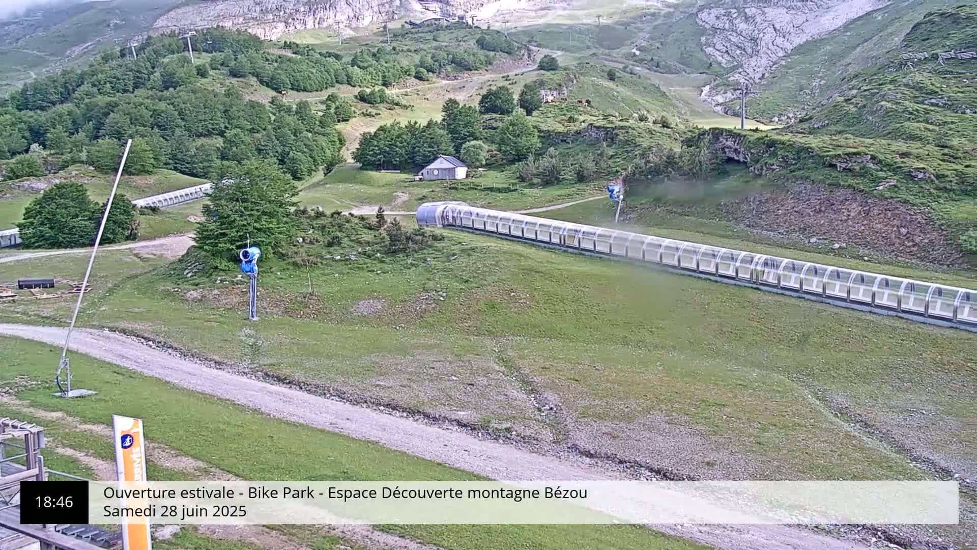 A long, covered mountainside slide is situated on a grassy hill, with a small building visible in the background under overcast skies.
