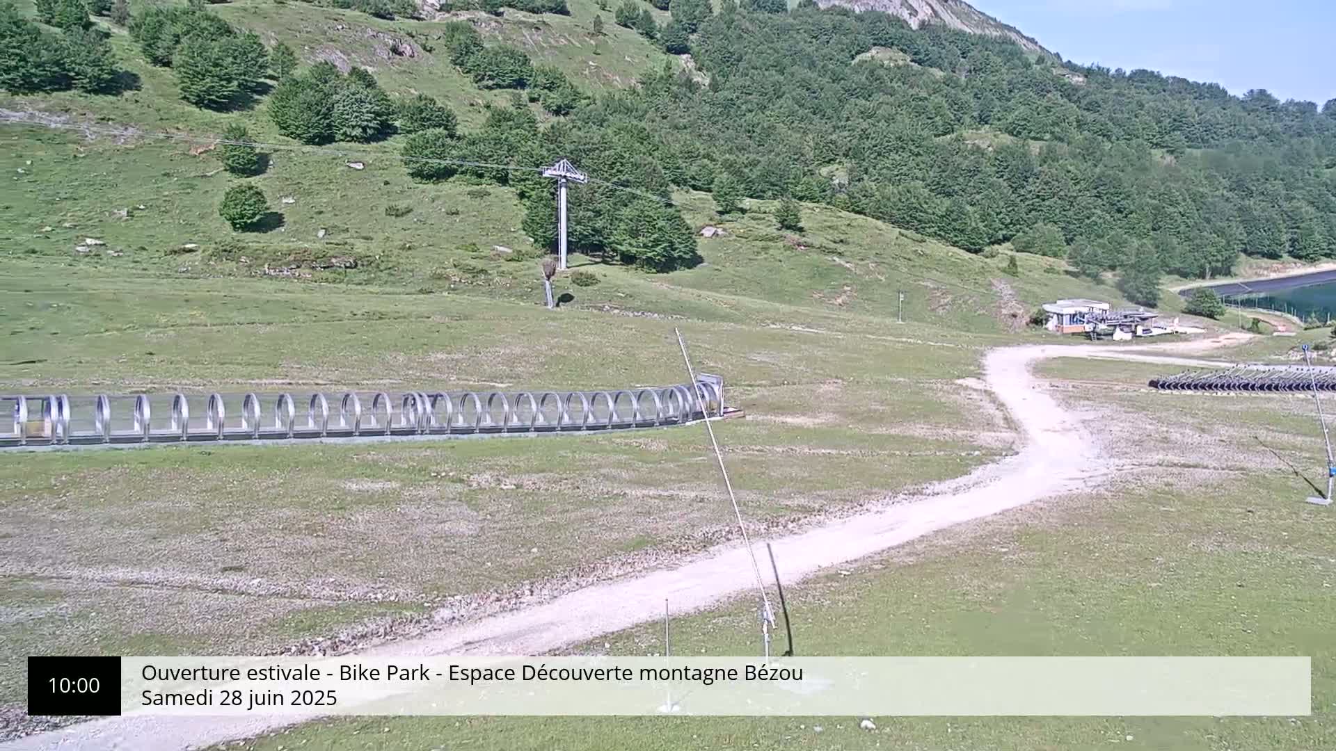 A mountainside bike park on a sunny day features a long, covered path, a chairlift, and a dirt road leading to a small building.