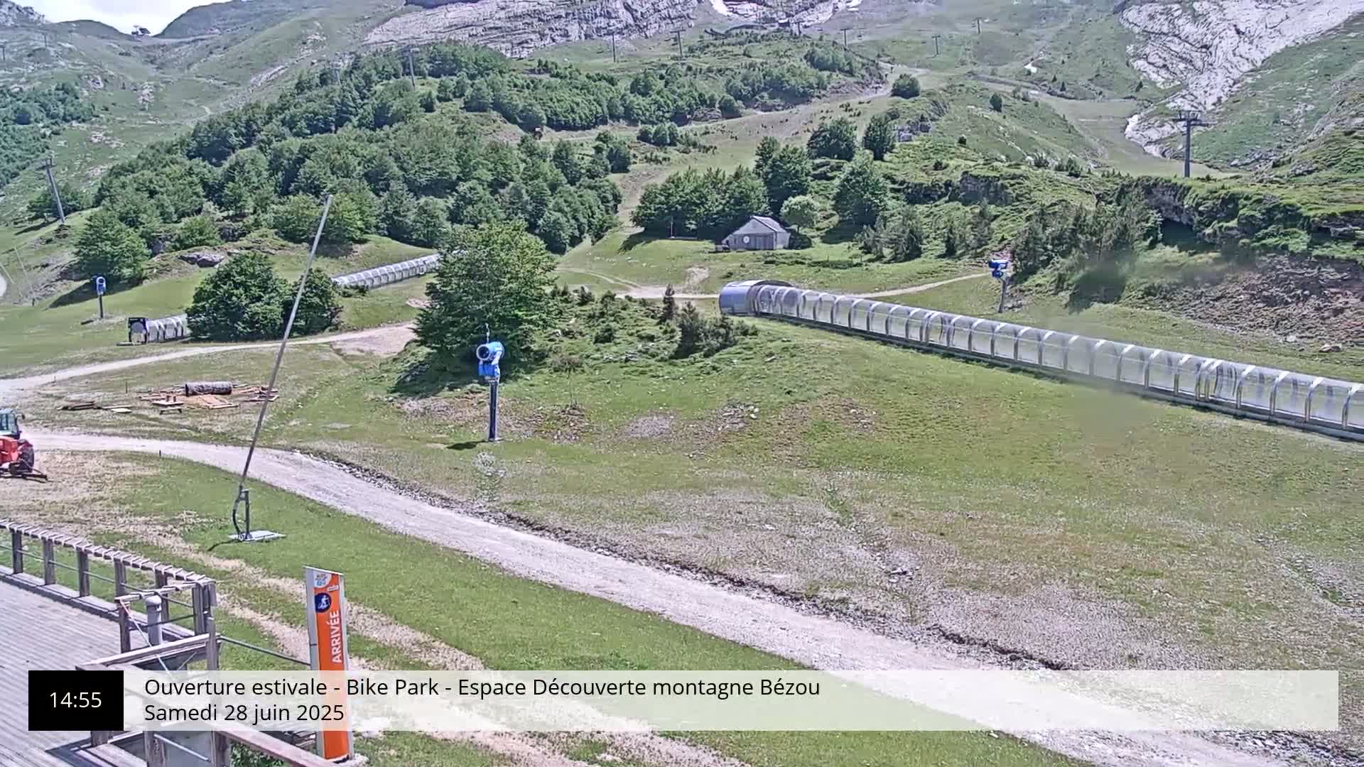 A mountain bike park with a long covered track, a small building, and a snow-making machine, all under sunny skies.