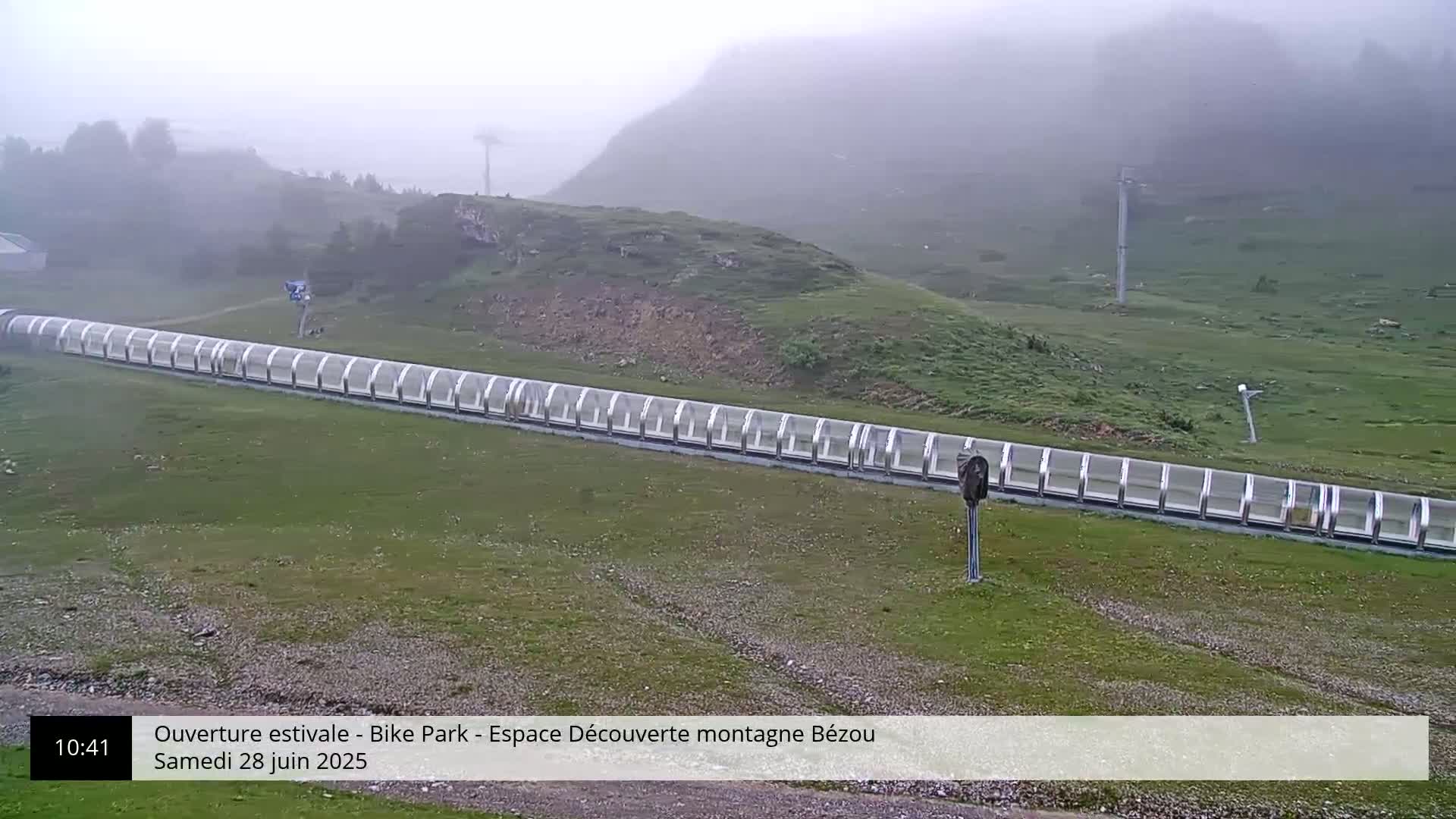 A long, covered conveyor belt-like structure sits on a grassy hillside in foggy conditions.