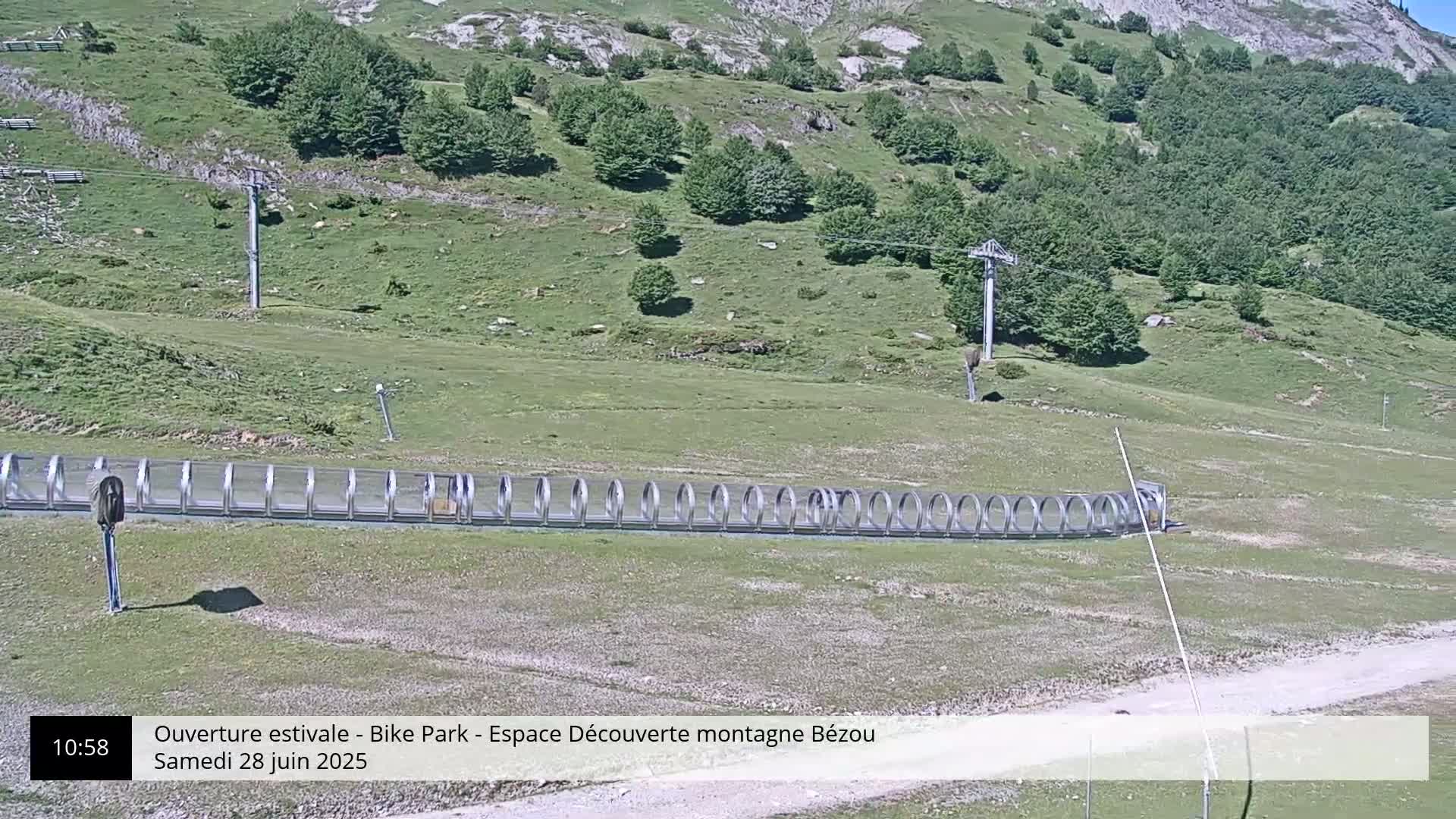 A mountainside with green grass and trees, a covered moving walkway, and a ski lift under a sunny sky.