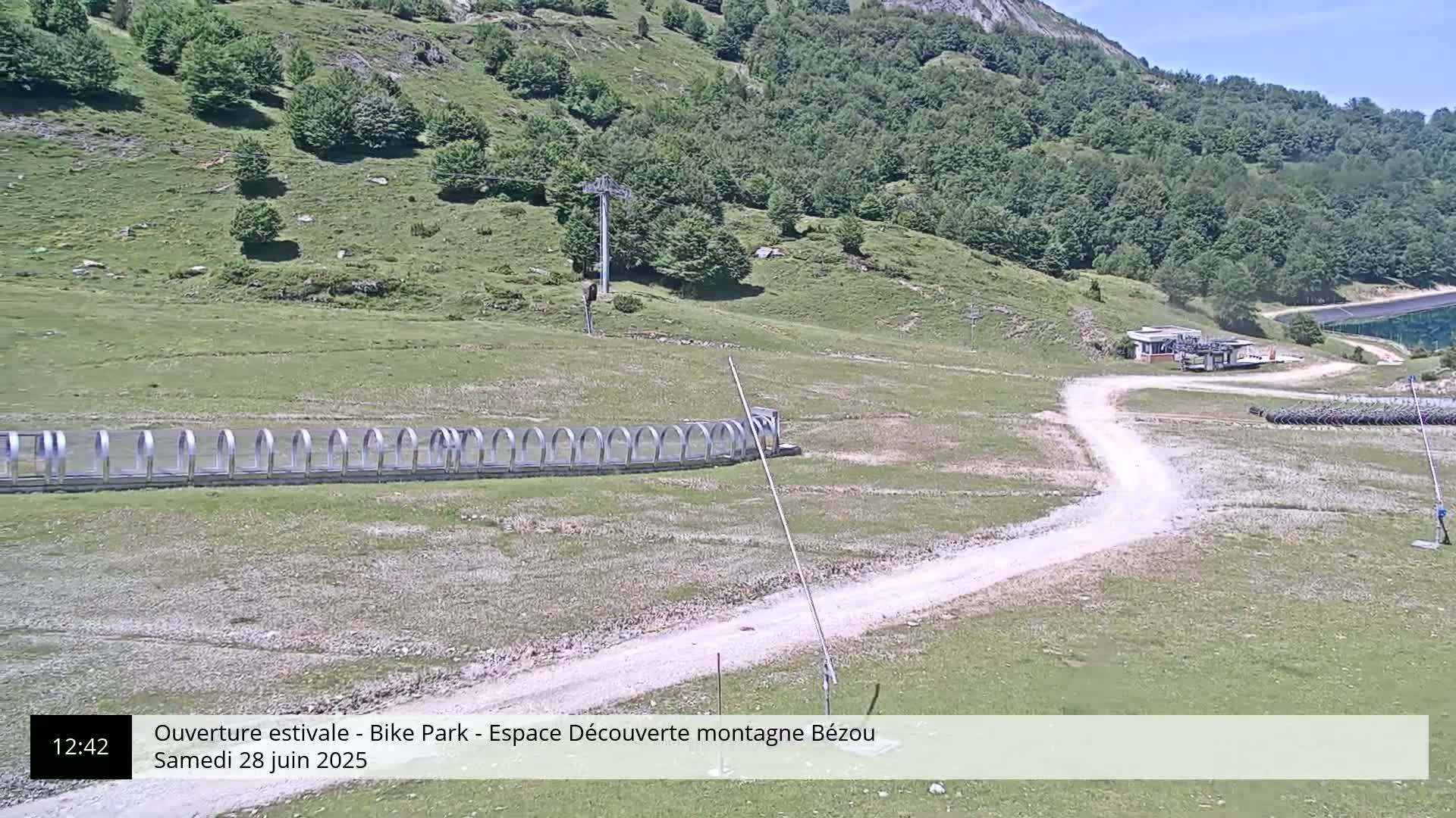 A mountaintop dirt road winds past a row of protective barriers, toward a small building and a chairlift against a backdrop of green hills under a sunny sky.