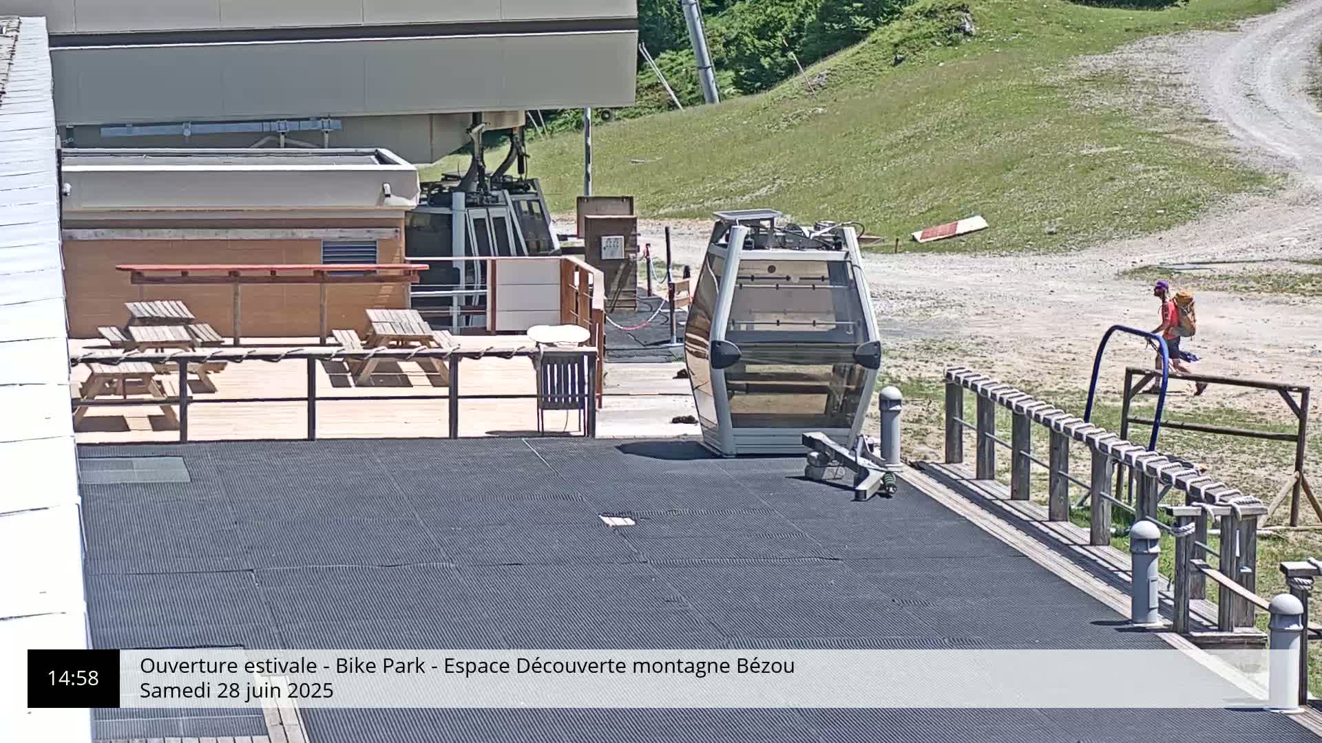 A mountaintop patio with picnic tables and a gondola car sits adjacent to a dirt path on a sunny day.