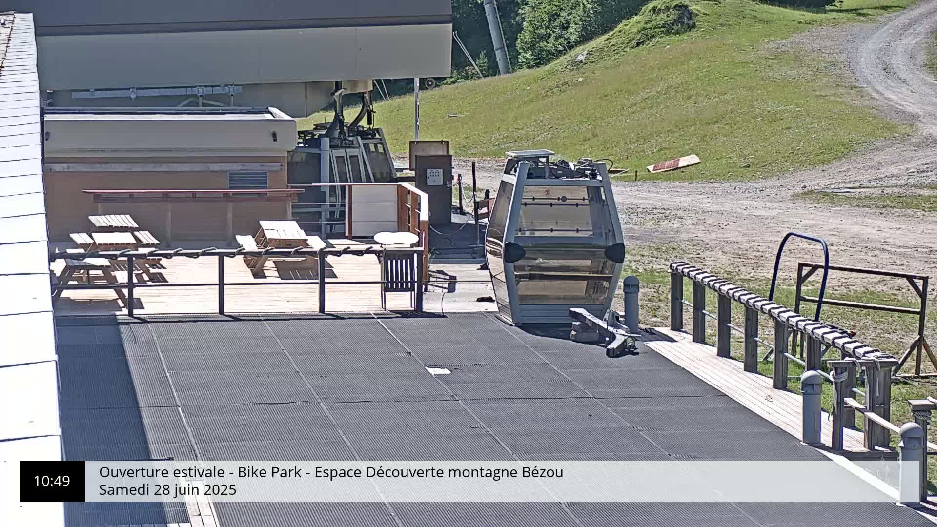 A cable car sits at a mountaintop station with picnic tables on a deck, under sunny conditions.