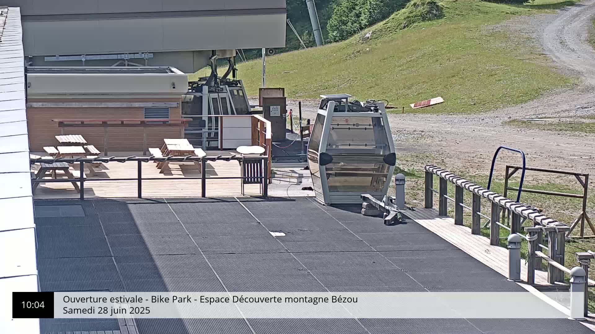 A sunny outdoor scene shows a chairlift gondola at a mountaintop station with picnic tables on a deck.