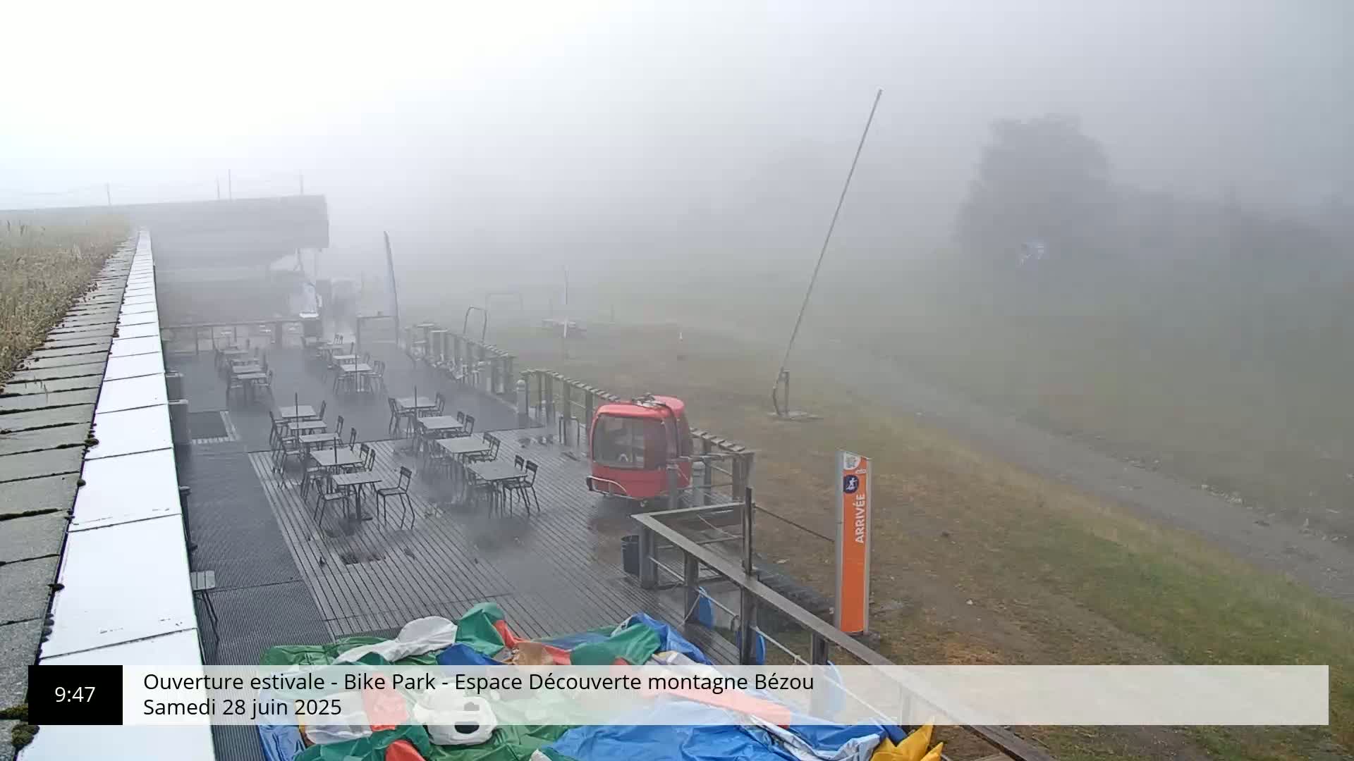 A foggy outdoor scene shows an empty outdoor restaurant with tables and chairs on a deck near a red cable car gondola.