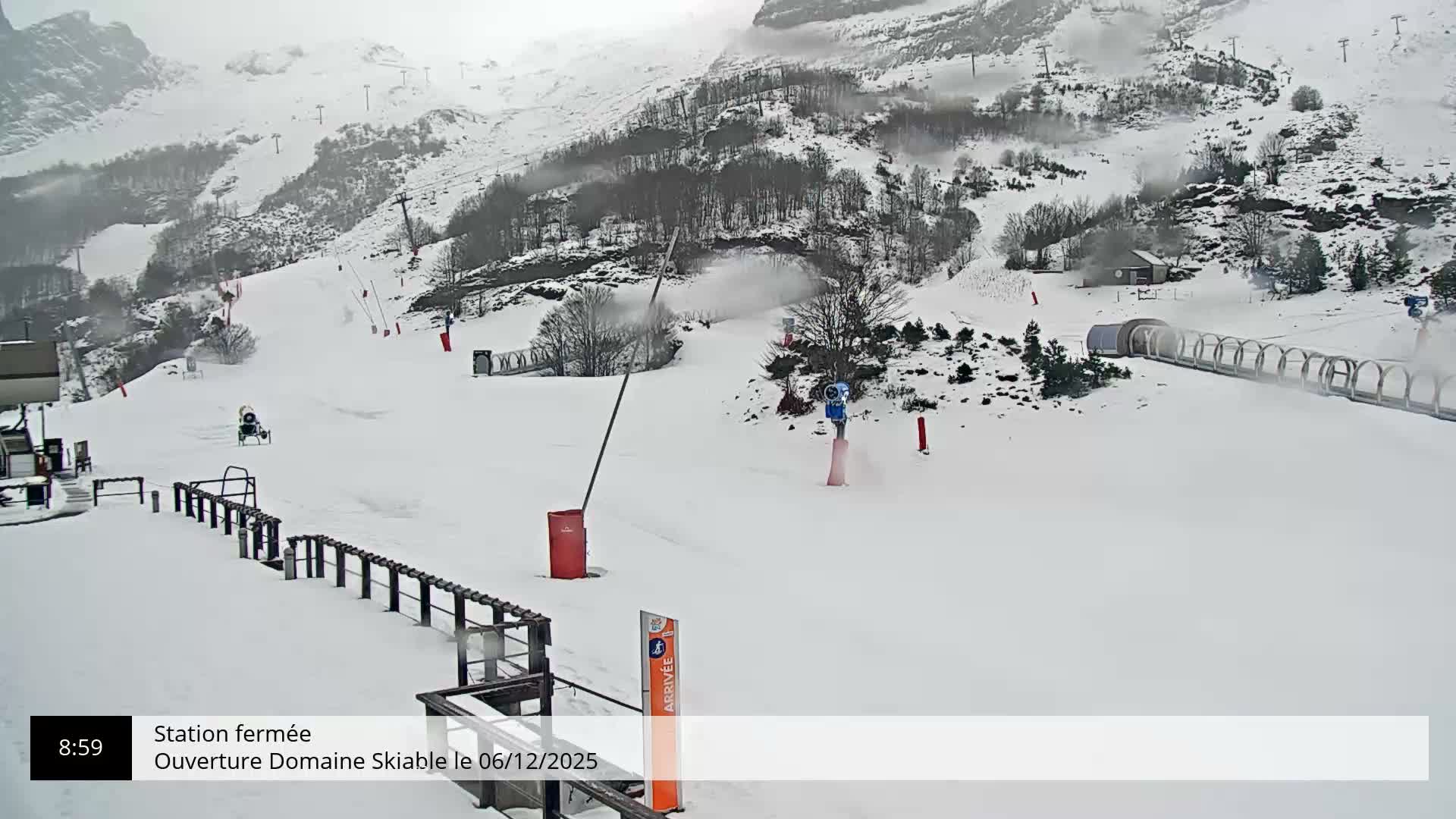 A snowy ski resort is depicted on an overcast day, with multiple snow cannons actively spraying snow onto the white slopes and surrounding hills dotted with trees and ski lifts.