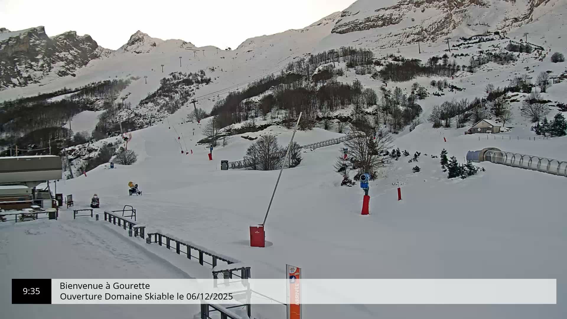 Under an overcast sky, a snow-covered mountain ski resort features wide slopes dotted with ski lifts, snow cannons, and a transparent tunnel, with a few small snow vehicles visible on the runs.
