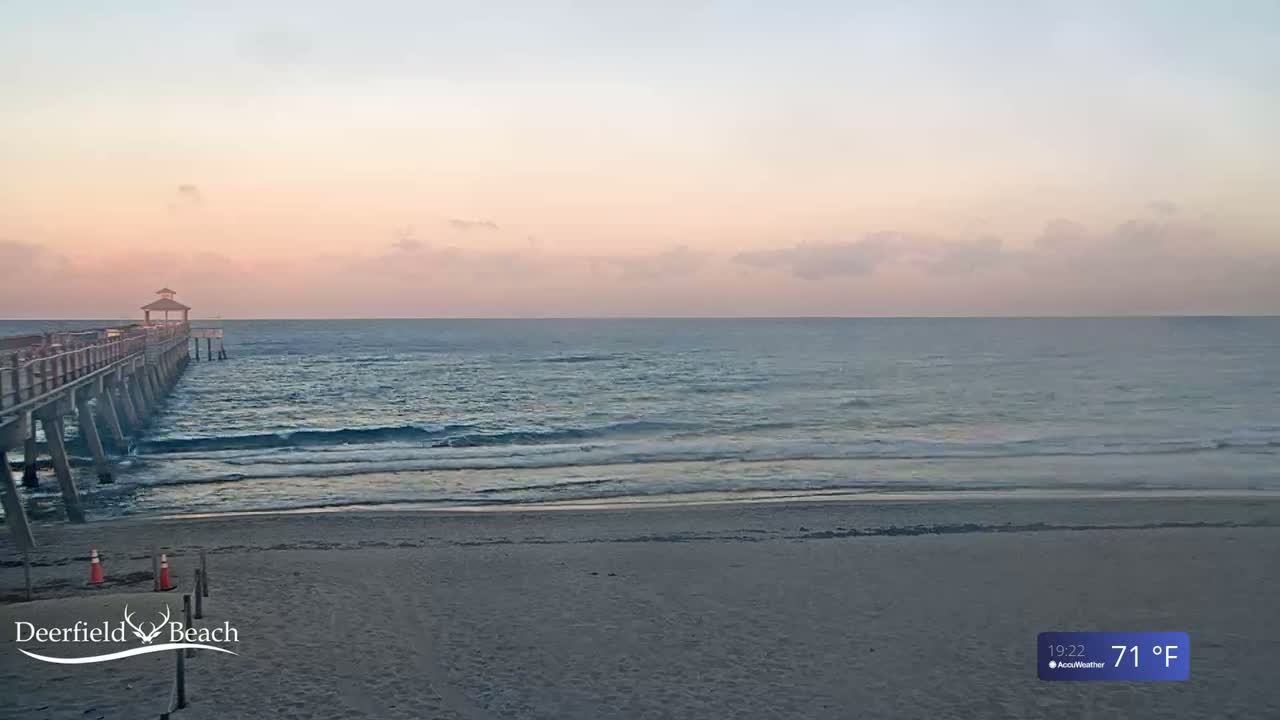 A long pier extends into a calm ocean under a pale pink and blue sunrise sky.