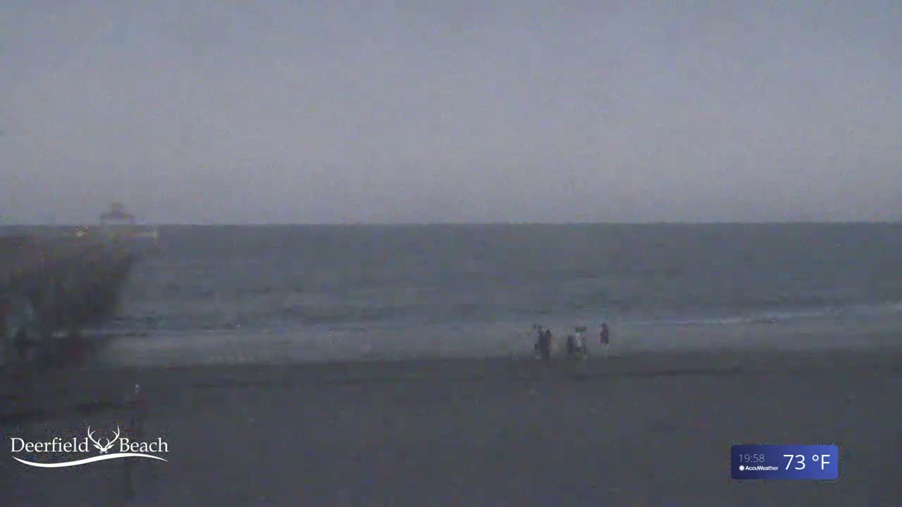 A hazy, overcast sky hangs over a dark beach with a few people walking near the water, which is relatively calm.