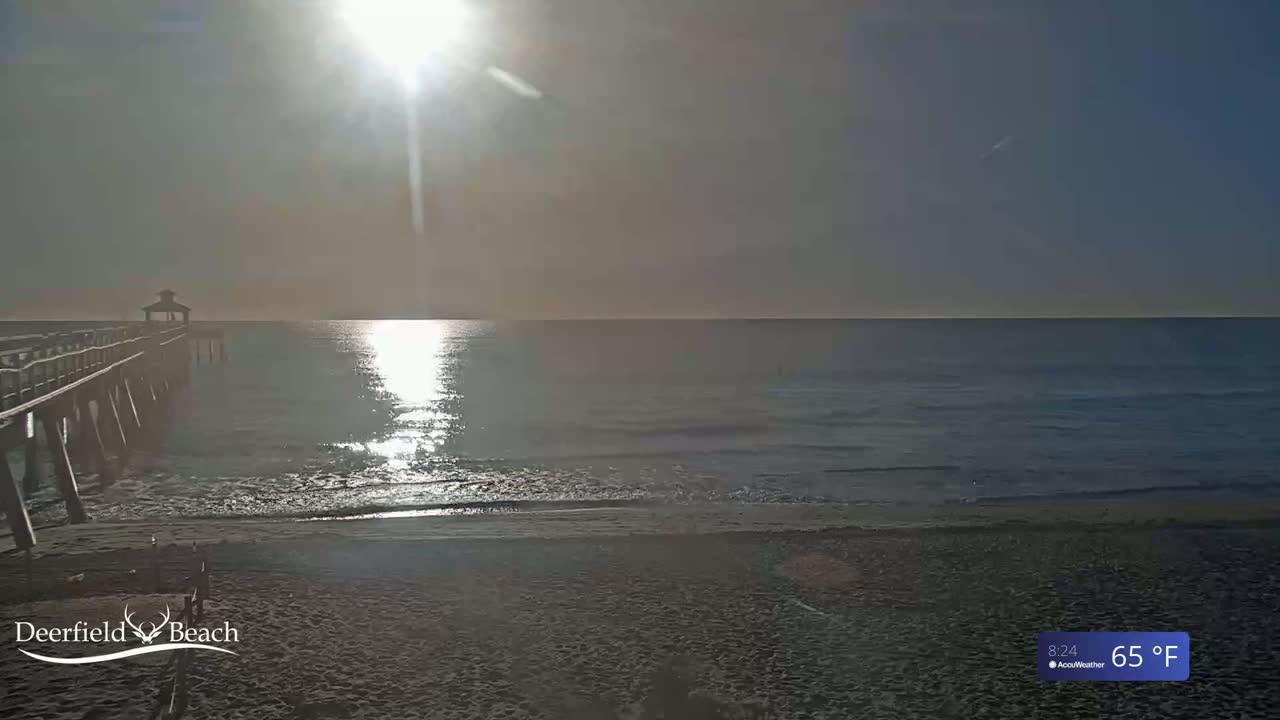 A long wooden pier extends into a calm ocean under a bright, hazy sun on a 65°F day.