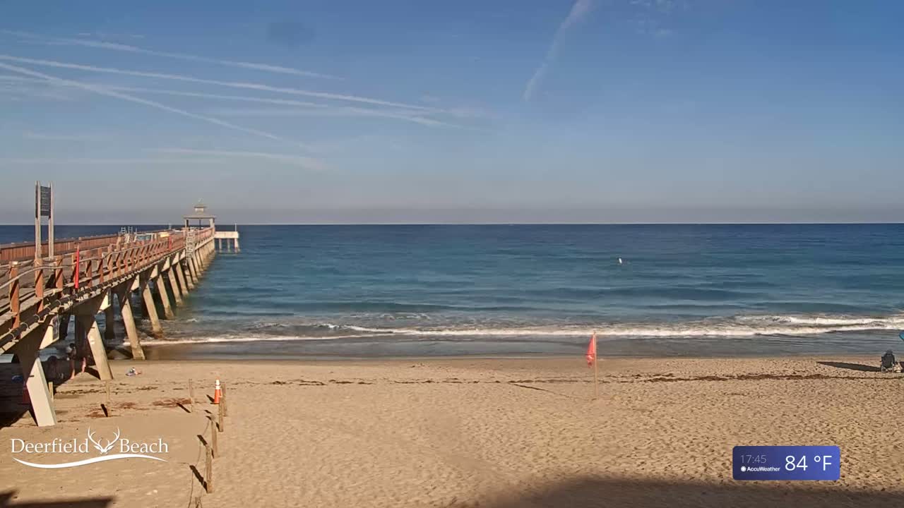 A long wooden pier extends over a sandy beach and calm, teal ocean under a clear blue sky.