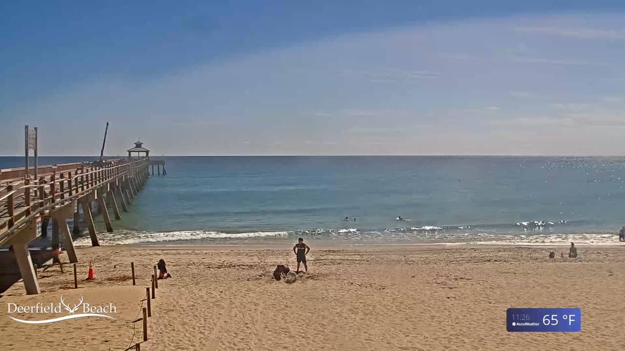 A long wooden pier extends over a sandy beach into a calm, light-blue ocean under a clear, sunny sky.