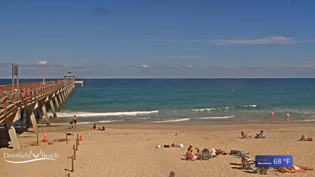 A long wooden pier extends over a sandy beach with calm, teal ocean waves under a clear blue sky.