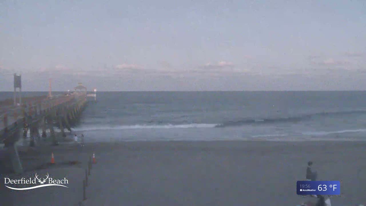 A long pier extends over a calm, grey ocean under a mostly cloudy, light grey sky.