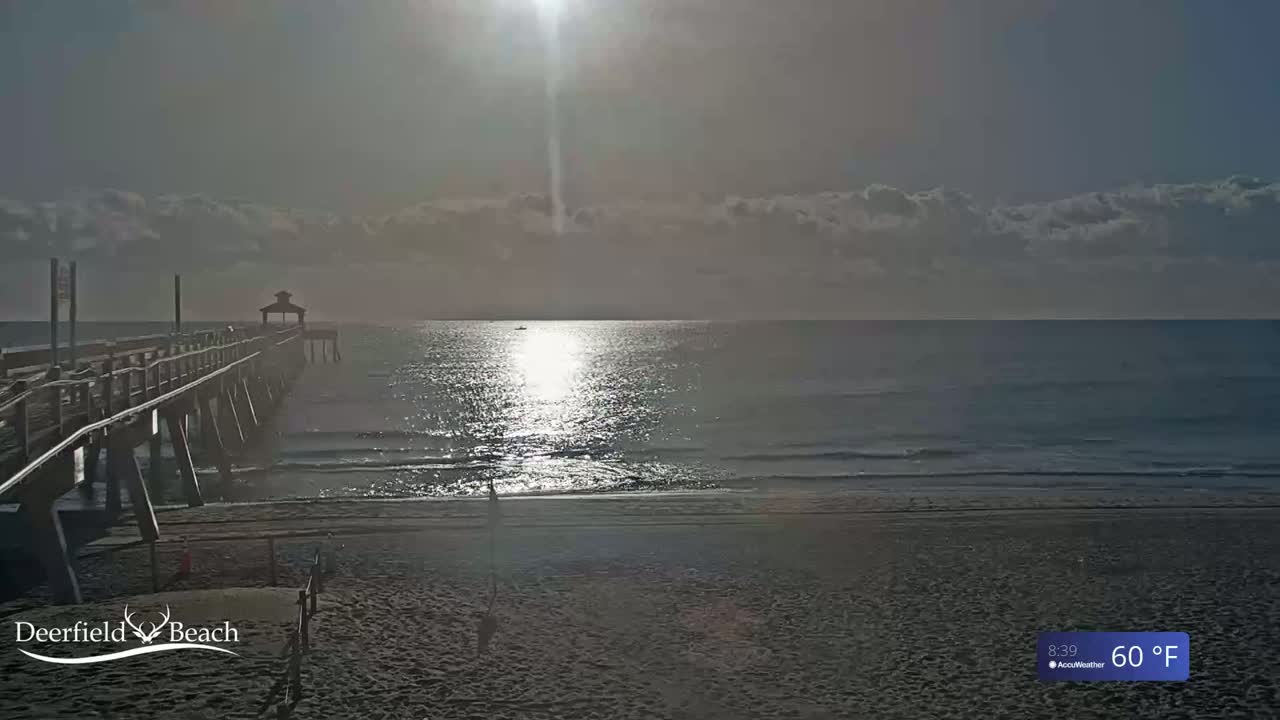 A long pier extends over a calm ocean under a mostly cloudy sky, with the sun shining brightly.