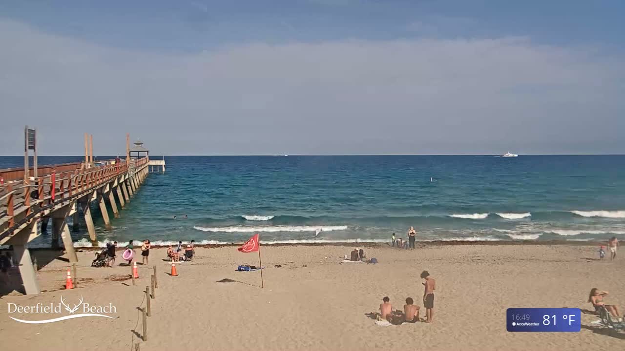 A long wooden pier extends over a sandy beach with calm ocean waves under a mostly sunny sky.