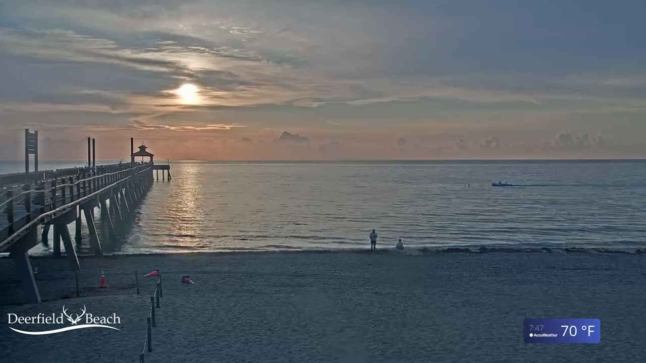 A long pier extends over calm ocean water at sunrise, with two people standing on a sandy beach in the foreground under partly cloudy skies, and a temperature of 70°F.