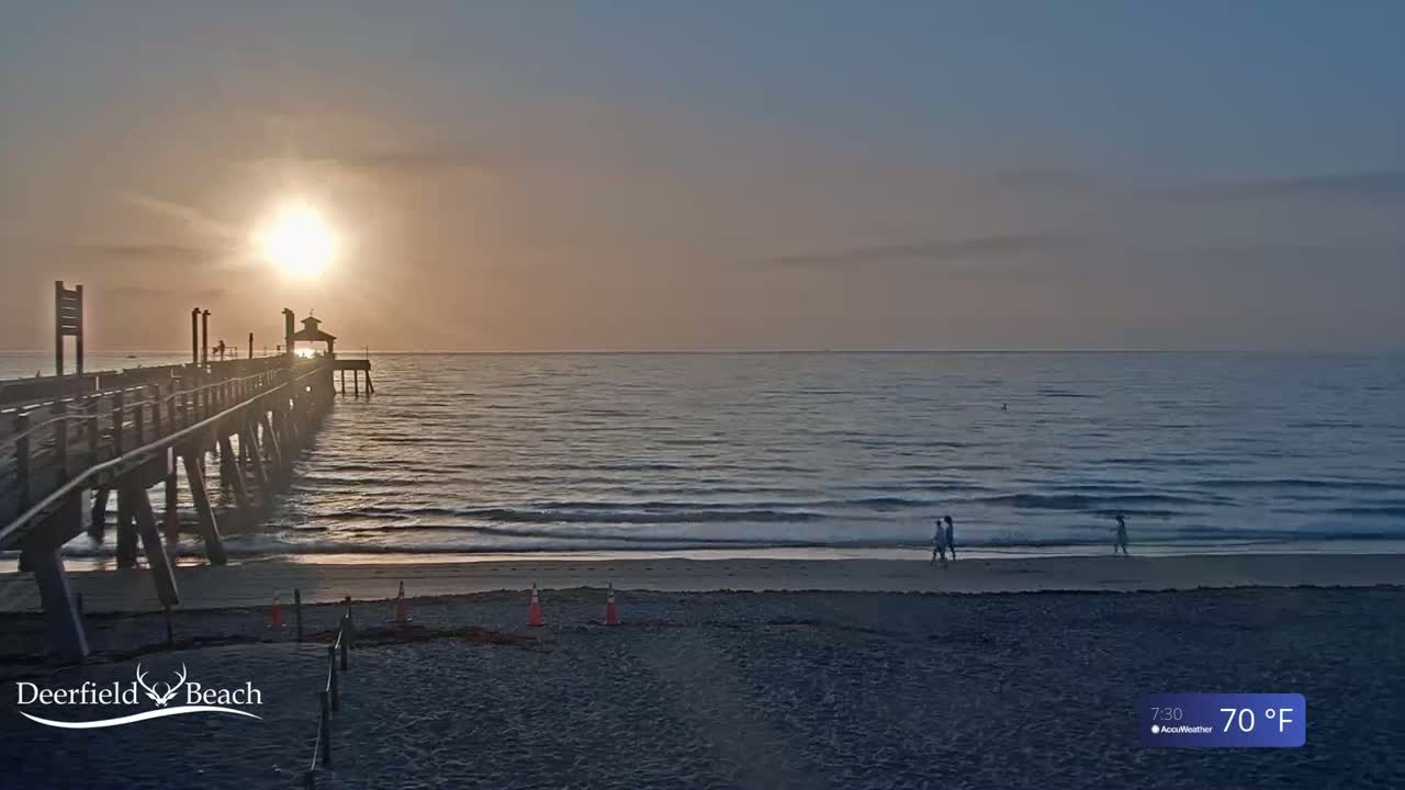 A long pier extends over a calm ocean at sunrise, with a few people walking on the beach in the soft light.