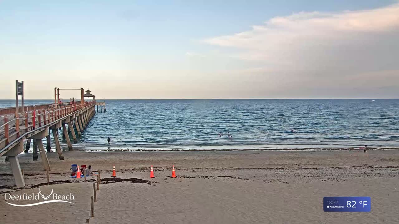 A sandy beach with a long pier extends into a calm, blue ocean under a partly cloudy sky.