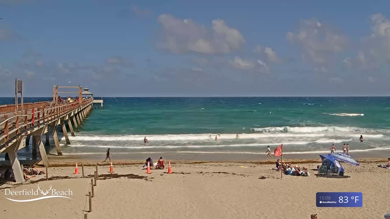 A long wooden pier extends over a sandy beach with people swimming and relaxing under a partly cloudy, sunny sky; the temperature is 83°F.