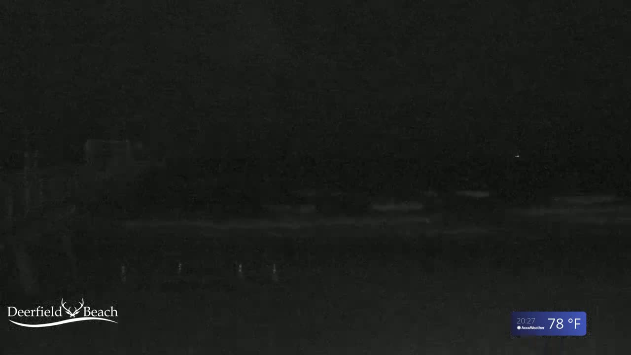 A dark, nighttime view of a beach shows a mostly black scene with a faint, lighter horizon line and a few small, distant lights.