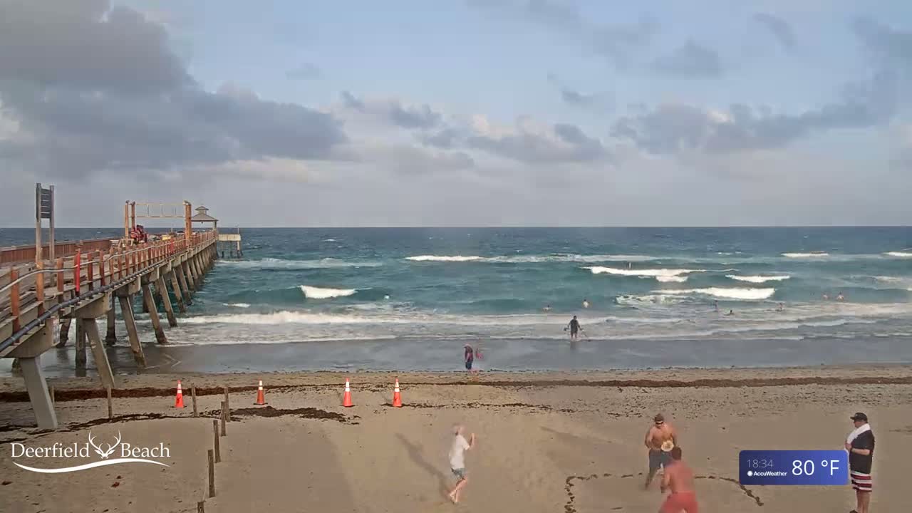 A mostly cloudy day shows a wooden pier extending over a sandy beach with moderate waves and several people on the sand and in the ocean.