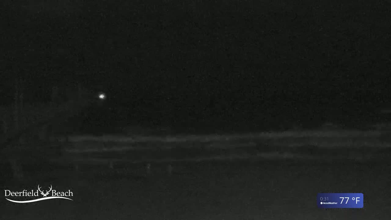 A nighttime, low-light image shows a dark beach under a clear sky with a single light source visible in the distance, at a temperature of 77°F.