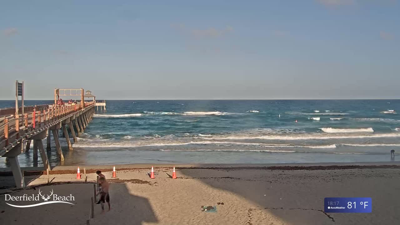 A long pier extends over a sandy beach with gentle waves rolling in under a clear, sunny sky.