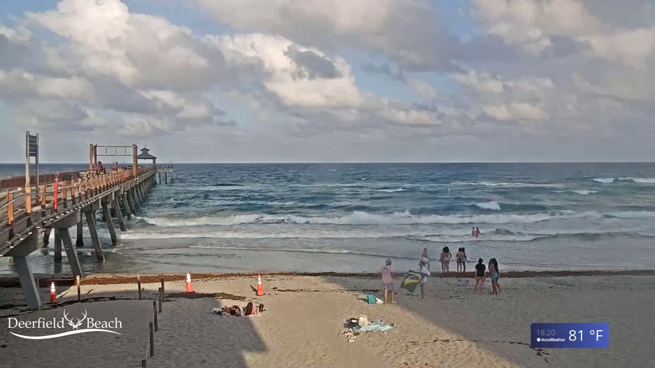 A long wooden pier extends over a sandy beach with a partly cloudy sky and moderate waves under a mostly cloudy sky.