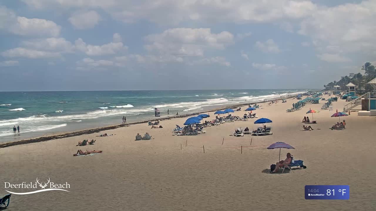 A sandy beach with people sunbathing under umbrellas, with a moderate surf under a partly cloudy sky.